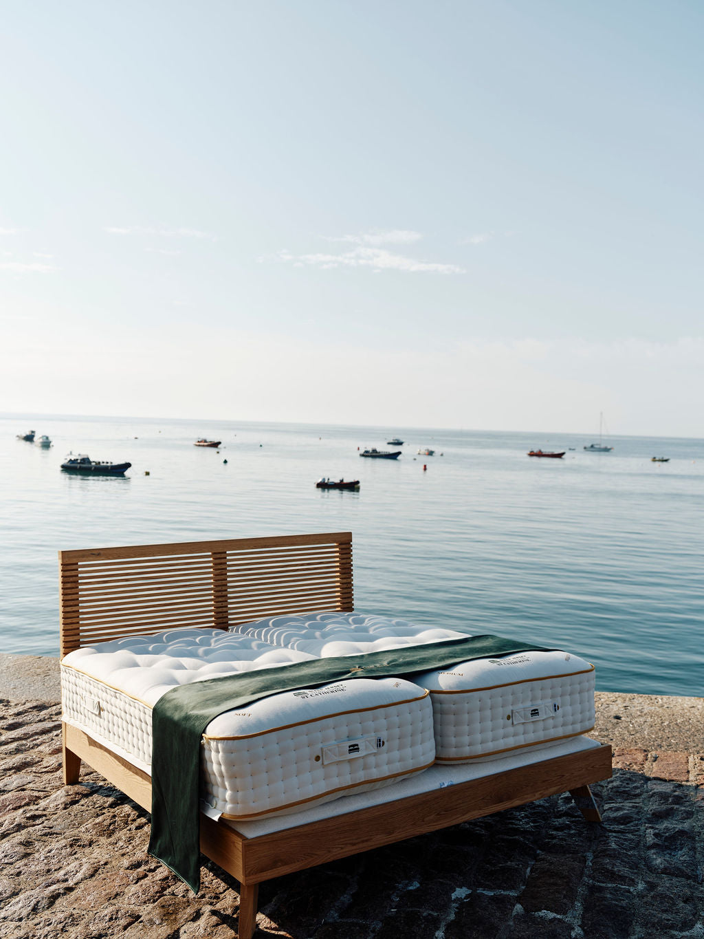 A wooden bed frame with two white mattresses and a green throw blanket, set outdoors on a stone surface overlooking a calm sea with small boats under a pale blue sky.